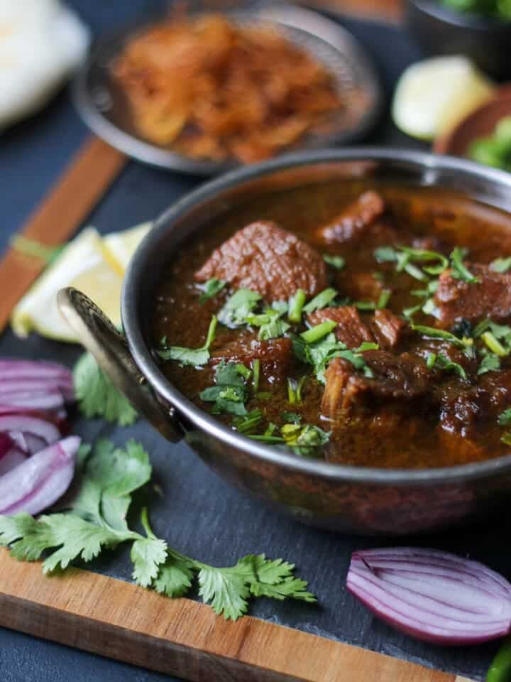 A bowl of spicy Chicken Kalia curry garnished with fresh cilantro, surrounded by sliced red onions, cilantro leaves, and other condiments on a wooden board.
