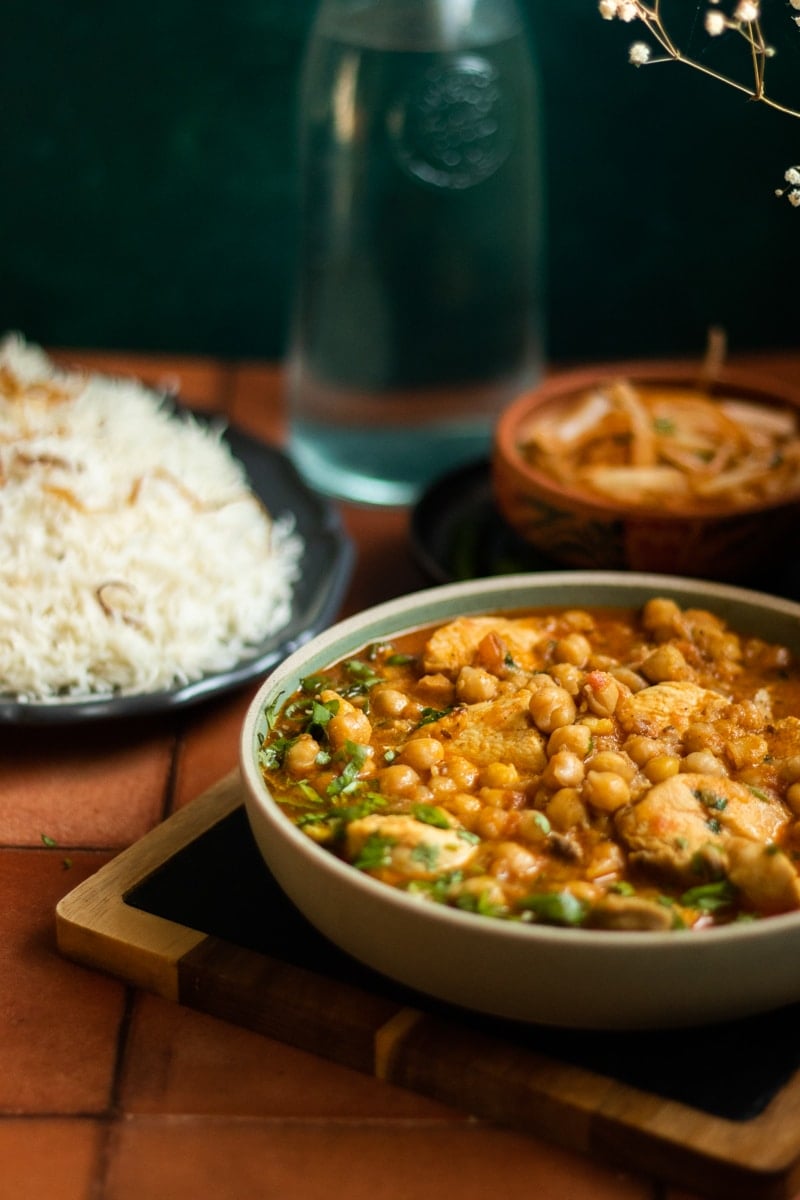¾ view of a dish of Murgh Cholay, a plate of rice in the background along with a bottle of water and sliced marinated onions.