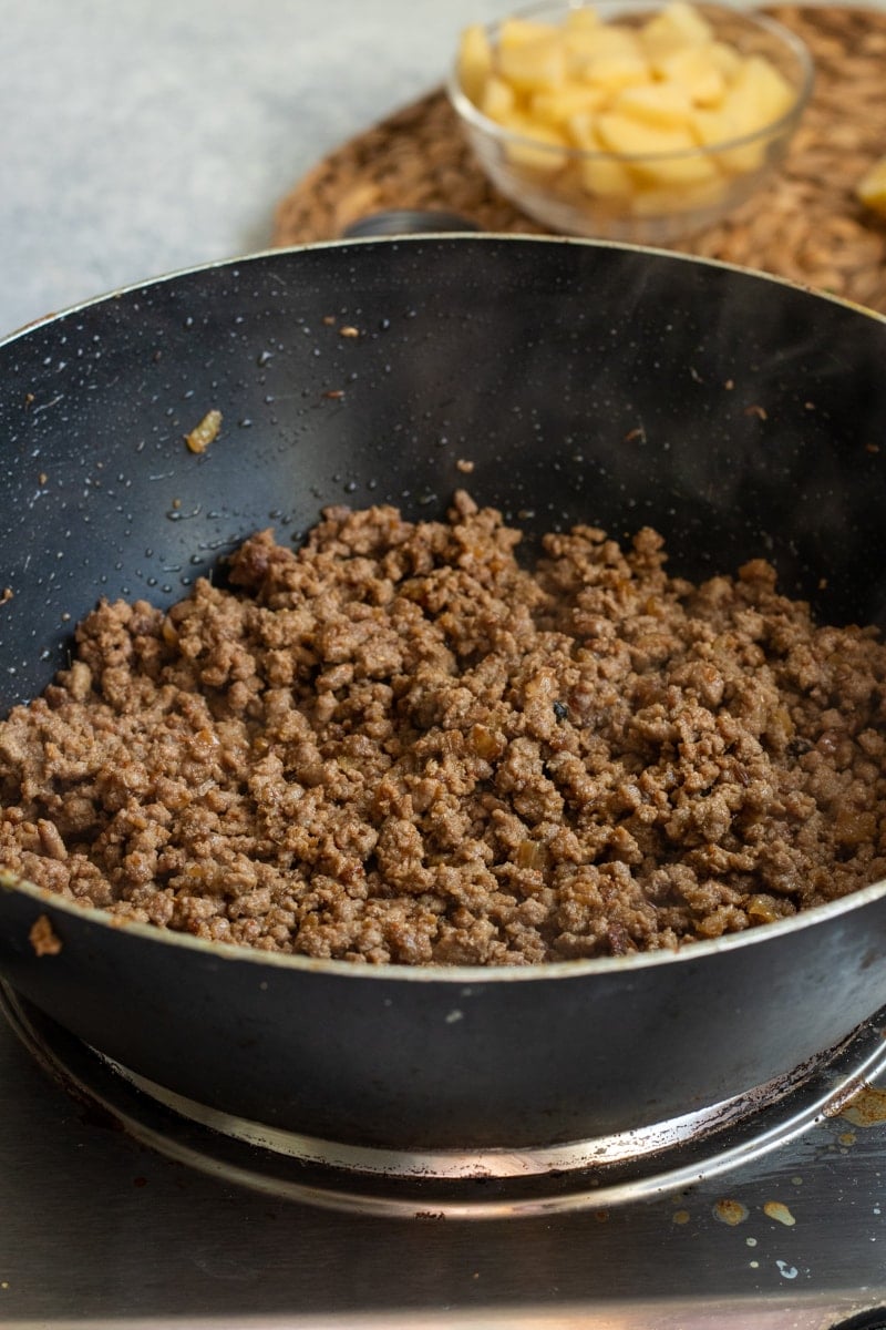 A black wok on a stovetop filled with cooked Qeema, steam rising; in the background, a glass bowl contains chopped potatoes on a woven mat, hinting at an Aloo Keema recipe in progress.