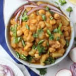 A bowl of aloo chana curry garnished with chopped onions, green chilies, and cilantro, surrounded by sliced radishes, green onions, lemon wedge, and a plate of rice on the side.