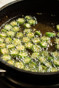 Sliced okra pieces frying in hot oil in a black skillet, with bubbles forming around the okra as it cooks—a delicious start to any Bhindi Masala Recipe.