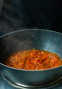 A black pan on a stove filled with chunky red tomato sauce simmering, steam rising against a dark background—perfect for starting your Bhindi Masala Recipe.