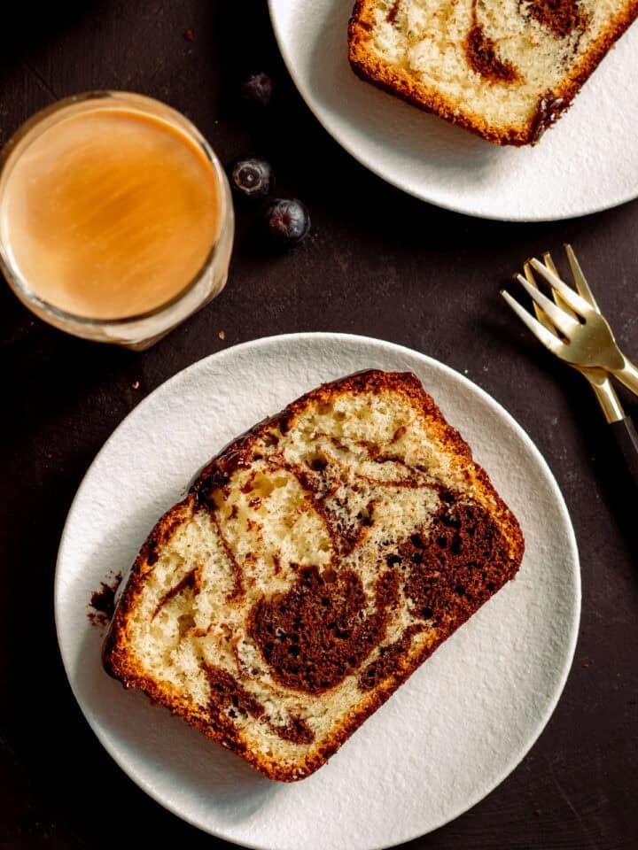 a slice of marble cake with oil on a plate, a cup of coffee and another half piece of marble cake partially visible