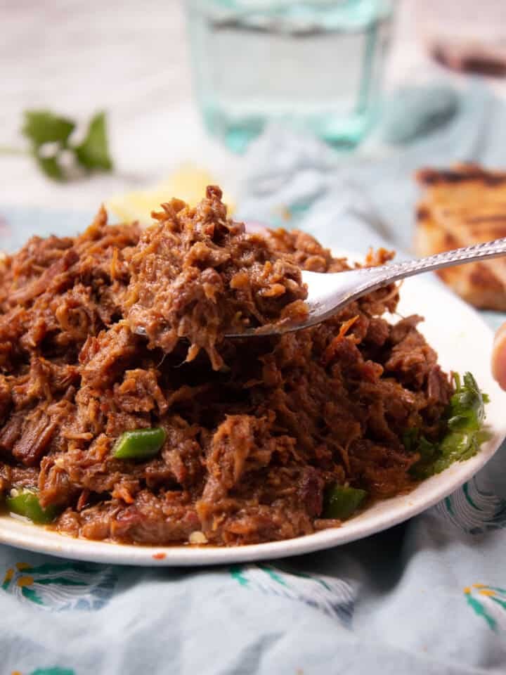 A close-up of a fork holding a portion of shredded Bhuna Gosht atop a plate, with green peppers visible underneath, set on a table with a blue cloth and glass of water in the background.