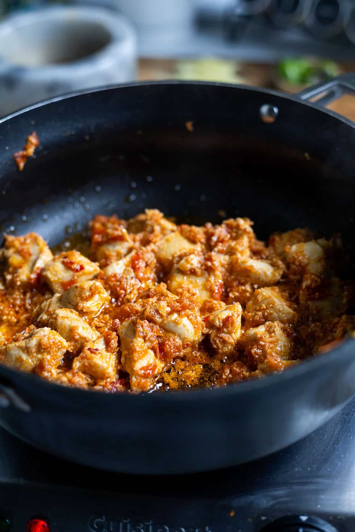 Chunks of chicken are cooking in a black pan, coated in a red, fragrant sauce-classic Karahi Chicken sizzling on the stovetop. The background is slightly blurred, revealing a warm kitchen setting.