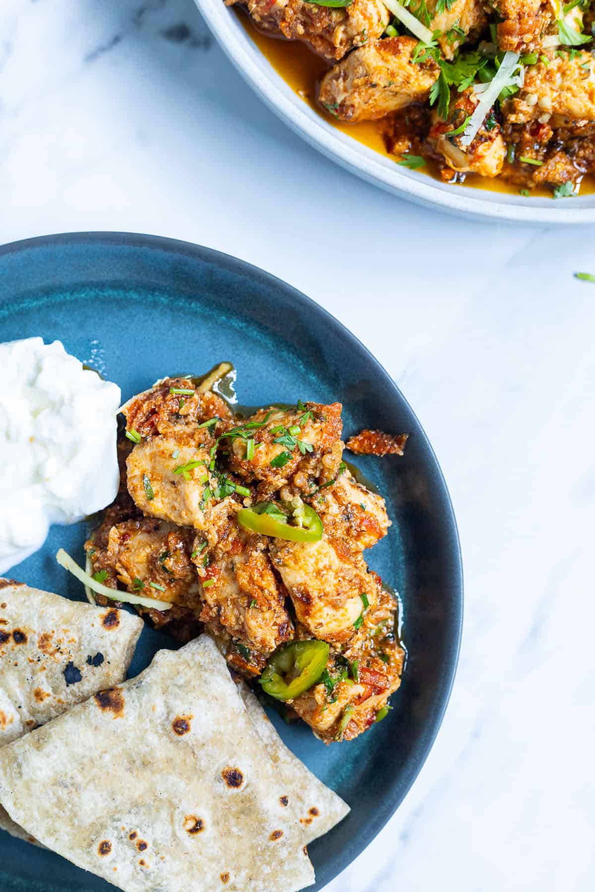 A blue plate with pieces of spiced Karahi Chicken curry garnished with green chilies and cilantro, served with two pieces of flatbread and a dollop of yogurt. Another dish of curry is partially visible above.