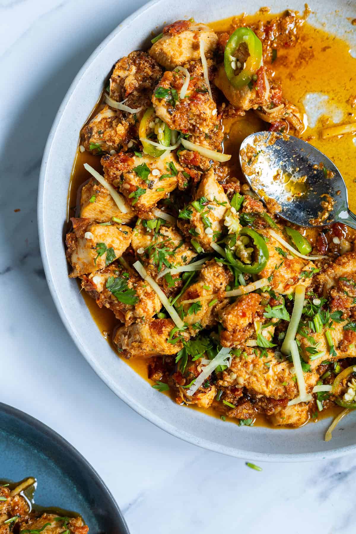 A plate of spicy Karahi Chicken curry garnished with chopped cilantro, sliced green chilies, and thin strips of ginger, with a serving spoon resting on the side.