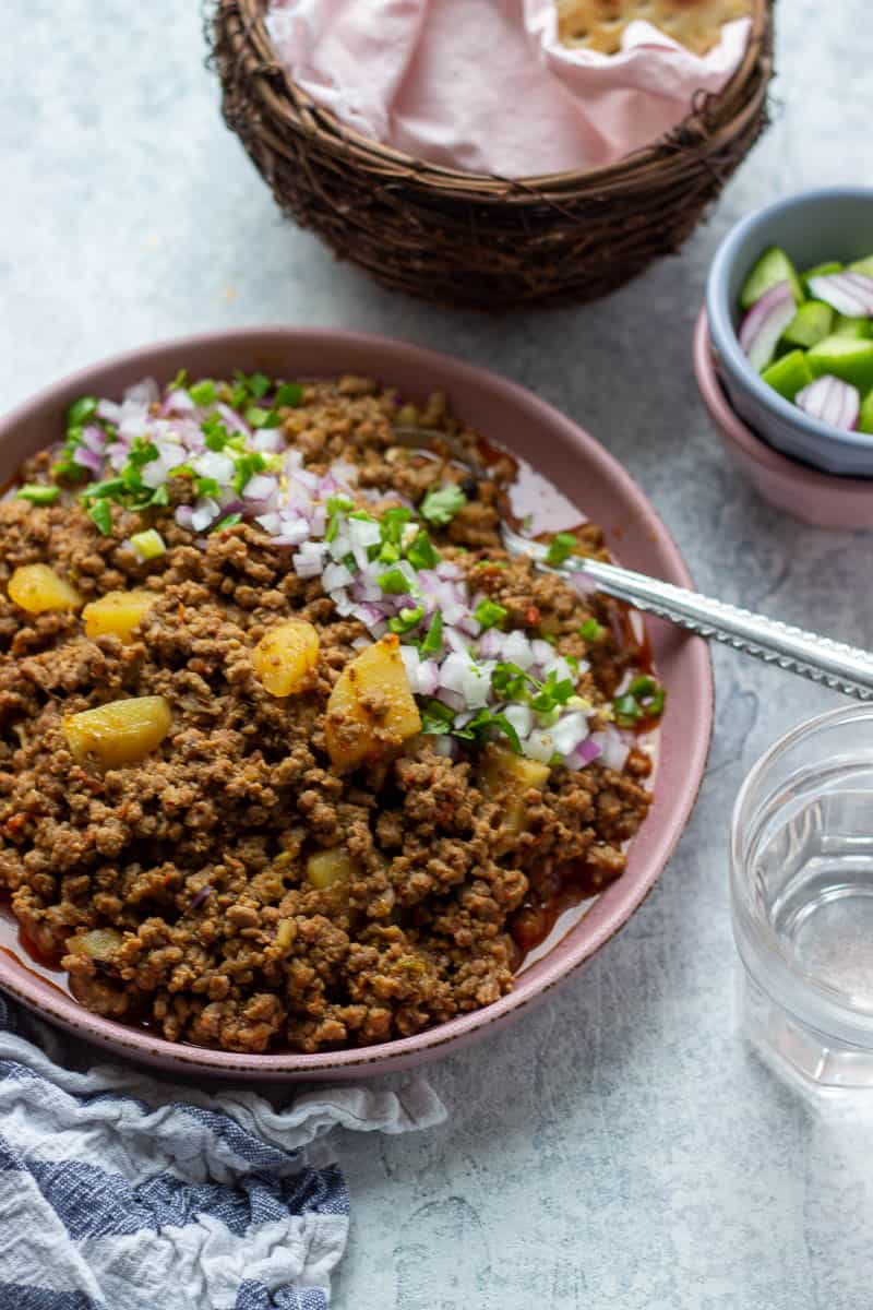 Aloo keema in a plate garnished with minced onions and chilies, some sheermal in a basket, chopped veg in a bowl