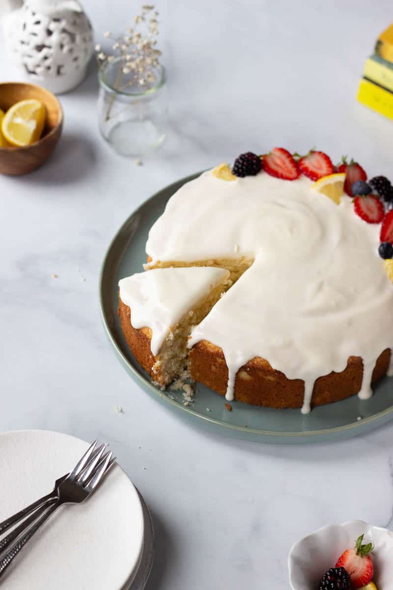 A lemon cake with cream cheese flaze with a slice partially pulled out. Plates, a little fruit and other yellow and white accents in the background