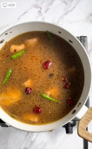 A pot of simmering Chicken Pilau soup with pieces of chicken, whole green chilies, and dried red chilies on a stove, photographed from above on a marble countertop.