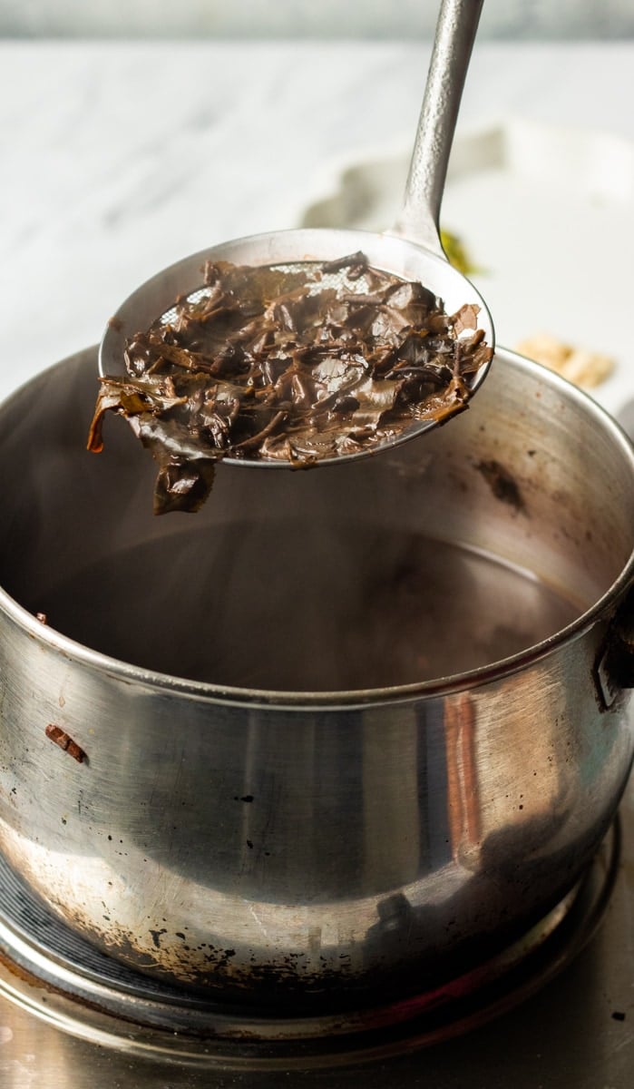 A metal ladle holds up a portion of cooked seaweed above a steaming pot of broth on a stovetop, reminiscent of the delicate preparation seen in traditional Pink tea.