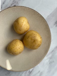 Three unpeeled yellow potatoes sit on a beige plate with a brown rim, placed on a white marble surface—perfect ingredients for making Aloo Palak.