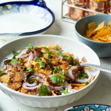 A bowl of Mixed Plate Chaat topped with sliced red onions, cilantro, green and brown chutneys, beside bowls of chickpeas, chips, and garnishes on a white table with decorative dishes in the background.