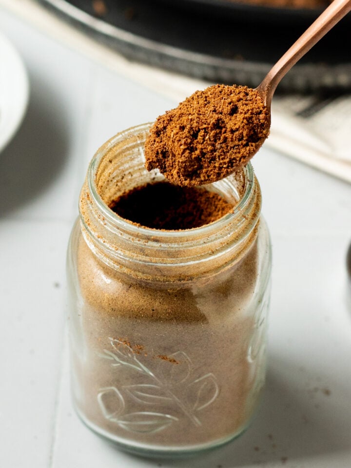 A close-up of a glass jar filled with ground coffee. A spoon is held above the jar, scooping out the coffee powder—reminiscent of how Pakistani Garam Masala is carefully measured. The jar sits on a white surface.