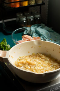 A white pan on a stovetop contains sautéed onions, the first step in making white chicken curry. In the background, a glass bowl holds raw chicken pieces, with spice jars, fresh green herbs, and a kitchen towel on the counter.