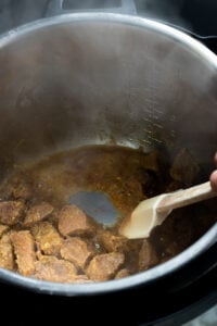 Chunks of beef being cooked for Tala hua gosht in a sauce inside a metal Instant Pot, with a white spatula stirring the meat. Steam rises from the pot, and measurement markings are visible on the pot’s inner wall.