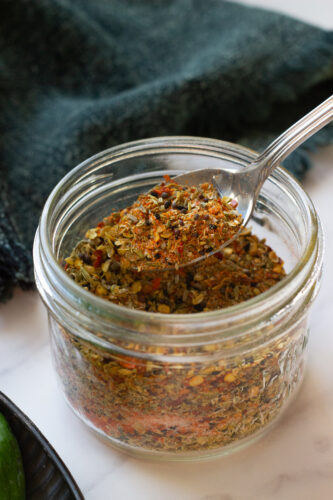 A close-up of a glass jar filled with colorful Achari Masala spice blend. A metal spoon scoops out the mixture, revealing dried herbs and crushed red pepper flakes. A dark cloth is in the background.