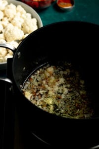 Chopped onions and spices sizzling in oil in a black pot, with a plate of raw cauliflower florets and some spices nearby—a classic start to Pakistani Aloo Gobi.
