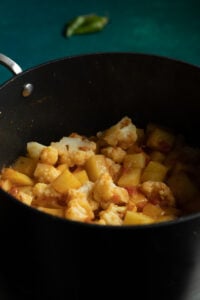 A black pot filled with Pakistani Aloo Gobi—cooked cauliflower and potato pieces in a spiced tomato sauce, with a blurred green chili in the background on a teal surface.
