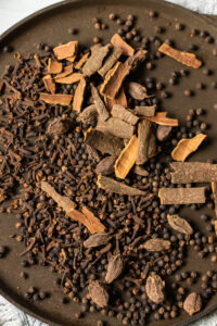A brown plate filled with whole spices—black peppercorns, dried cloves, cinnamon sticks, and large black cardamom pods—perfect for creating authentic Pakistani Garam Masala, all arranged in a loose pile.