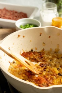 Onions sautéing in a white pan with spices and a wooden spoon; green peppers, raw Chicken Keema, and a jar of liquid are in the background.