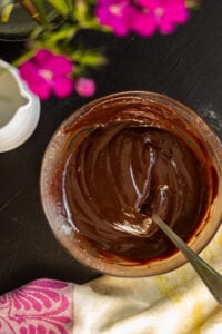 A bowl of smooth, glossy chocolate ganache with a spoon in it, placed on a dark surface near a pink and yellow patterned cloth and blurred pink flowers—perfect for topping your favorite marble cake with oil.
