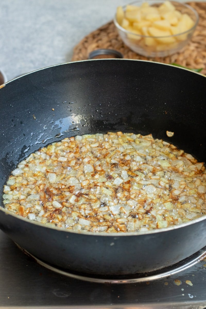 Chopped onions are being sautéed and browned in oil in a black frying pan on a stovetop,