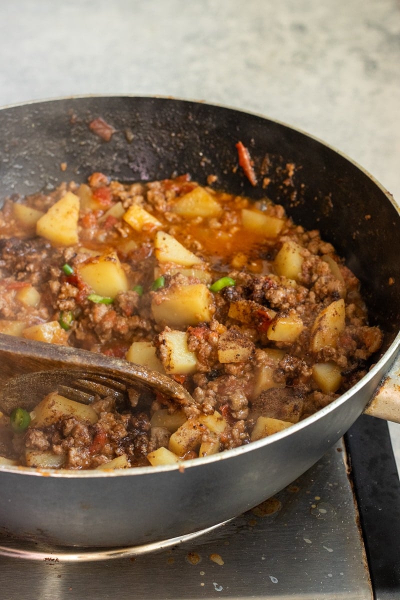 Ground beef and diced potatoes simmer in a tomato-based sauce inside a deep skillet, creating a classic Aloo Keema. A wooden spoon stirs the mixture as steam rises from this hot, hearty qeema dish.
