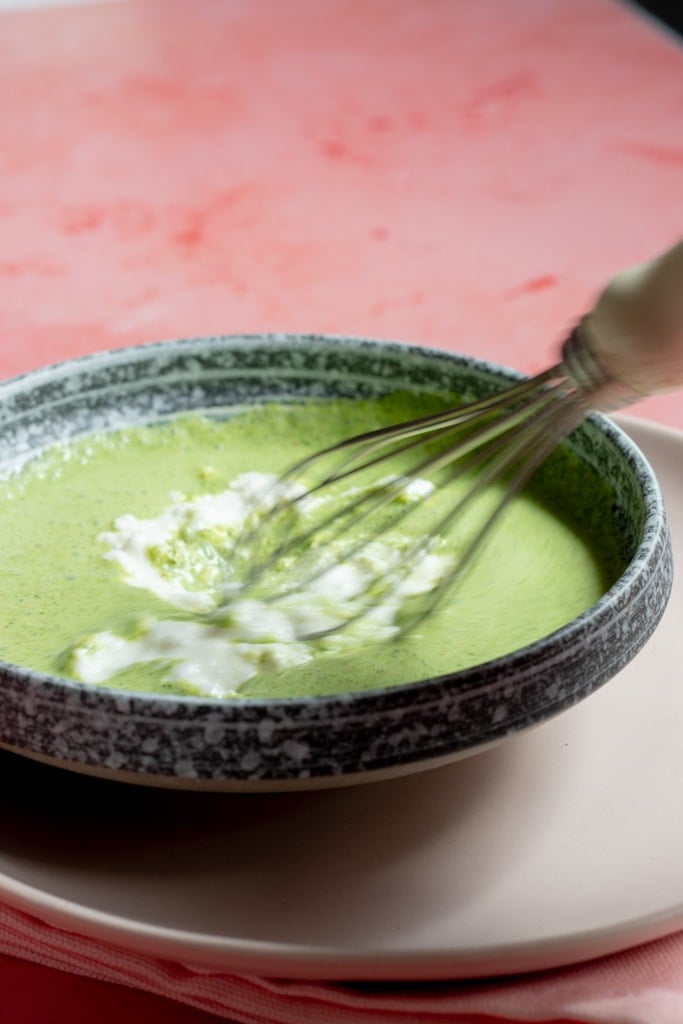 A whisk mixing a green creamy Pudina Raita with white chunks in a gray bowl on a white plate, set on a pink surface.