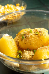 A glass bowl filled with whole, peeled boiled potatoes seasoned with ground spices, chopped green chilies, and black pepper—perfect for preparing Aloo Cutlets or following your favorite Potato Cutlets Recipe. A blurred bowl of corn kernels sits in the background.