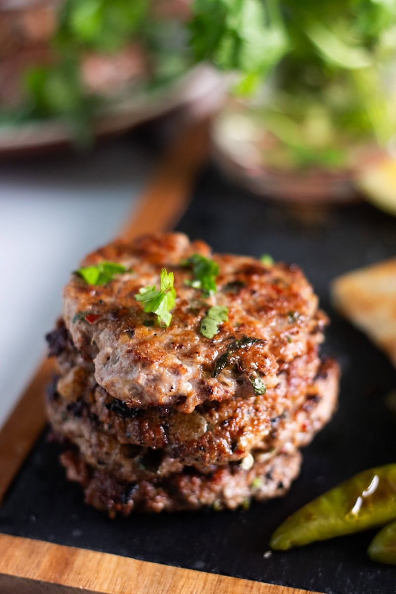a stack of chicken chapli kabab on a board, blistered green chilies in the corner 